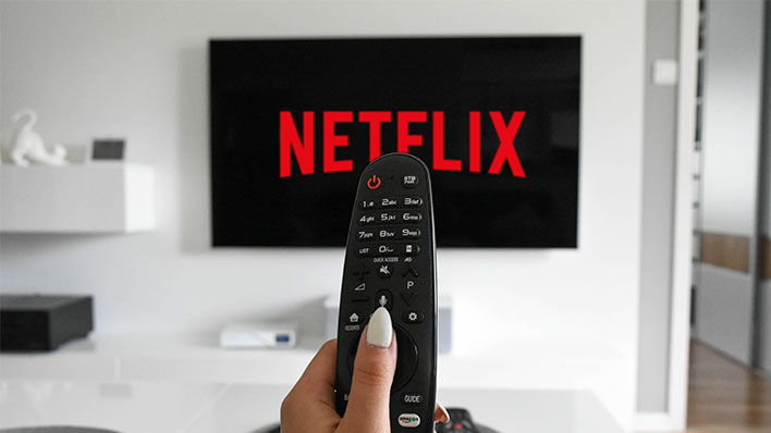 Hand pointing a remote control at a TV with "Netflix" displayed on it.