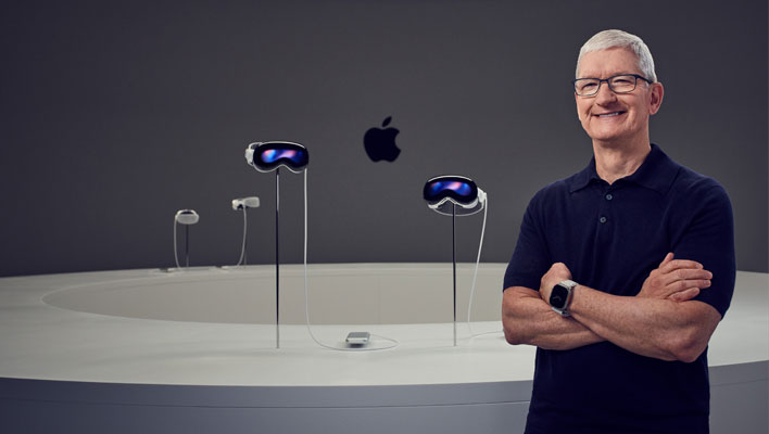 Apple CEO Tim Cook with folded arms and smiling standing in front of several Vision Pro headsets.