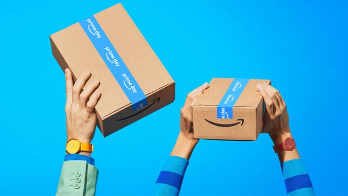 Arms hold up a pair of Amazon Prime Day boxes on a blue background.