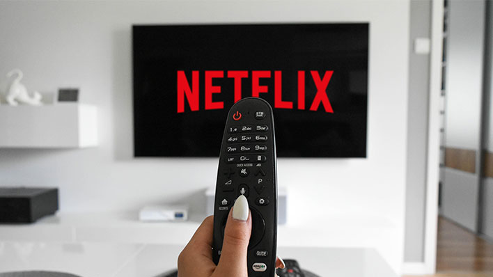Hand holding a remote in front of a TV with "Netflix" on the display.