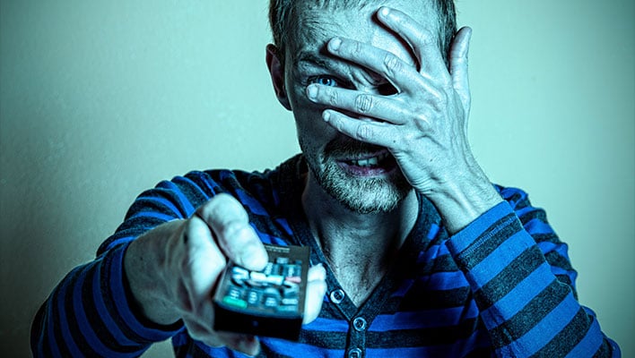 Man in a blue and black striped shirt covering his face with one hand and holding out a remote control in the other.