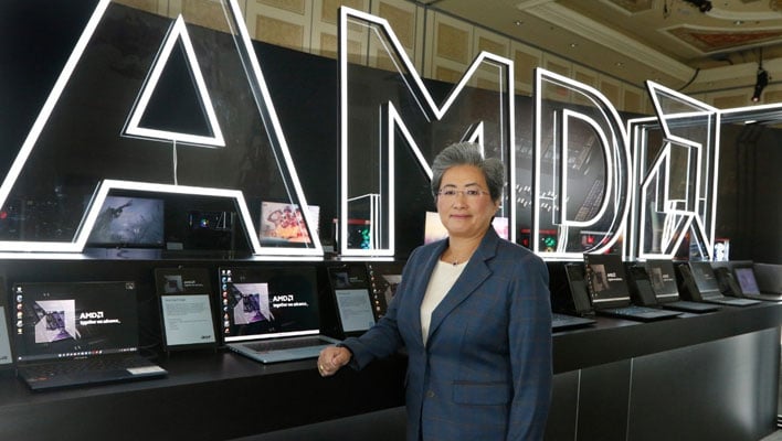 AMD CEO Dr. Lisa Su standing in front of a row of laptops and a big AMD sign.