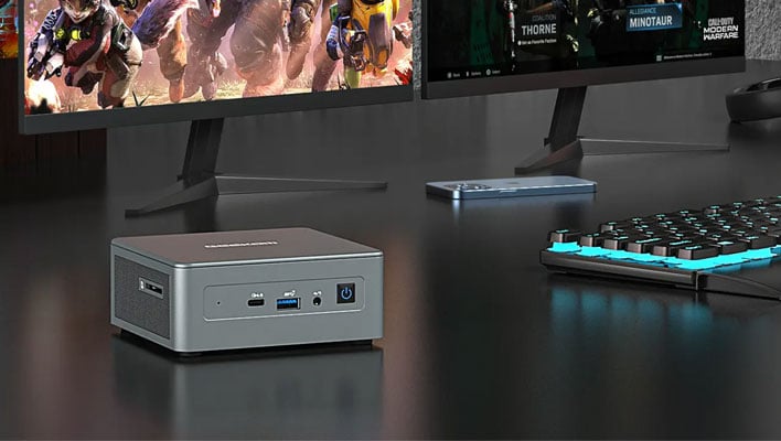 Geekcom mini PC on a desk next to two monitors and a keyboard.