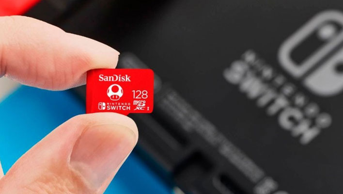Fingers holding a licensed 128GB SanDisk microSD card (red, with Mushroom) in front of a blurry Switch console.