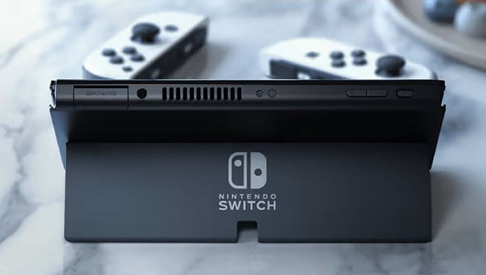 Rear view of the Nintendo Switch OLED on its kickstand, with two white Joy-Con controllers in front of it, on a table..
