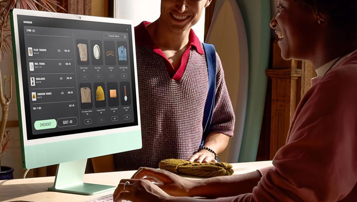 Person using an iMac on a desk. Another person is watching and smiling in the background.