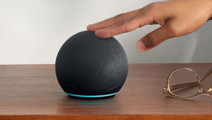 Amazon Echo Dot (5th Gen) on a nightstand. A hand is placed on top.